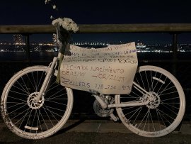 Ghost bike installed 2/26/2023 White painted ghostbike attached to a railing with flowers on the handlebar, a hand written sign in Spanish attached to it, photo taken at night, lights of NJ across the Hudson River in the background.