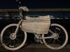 Ghost bike installed 2/26/2023 White painted ghostbike attached to a railing with flowers on the handlebar, a hand written sign in Spanish attached to it, photo taken at night, lights of NJ across the Hudson River in the background.